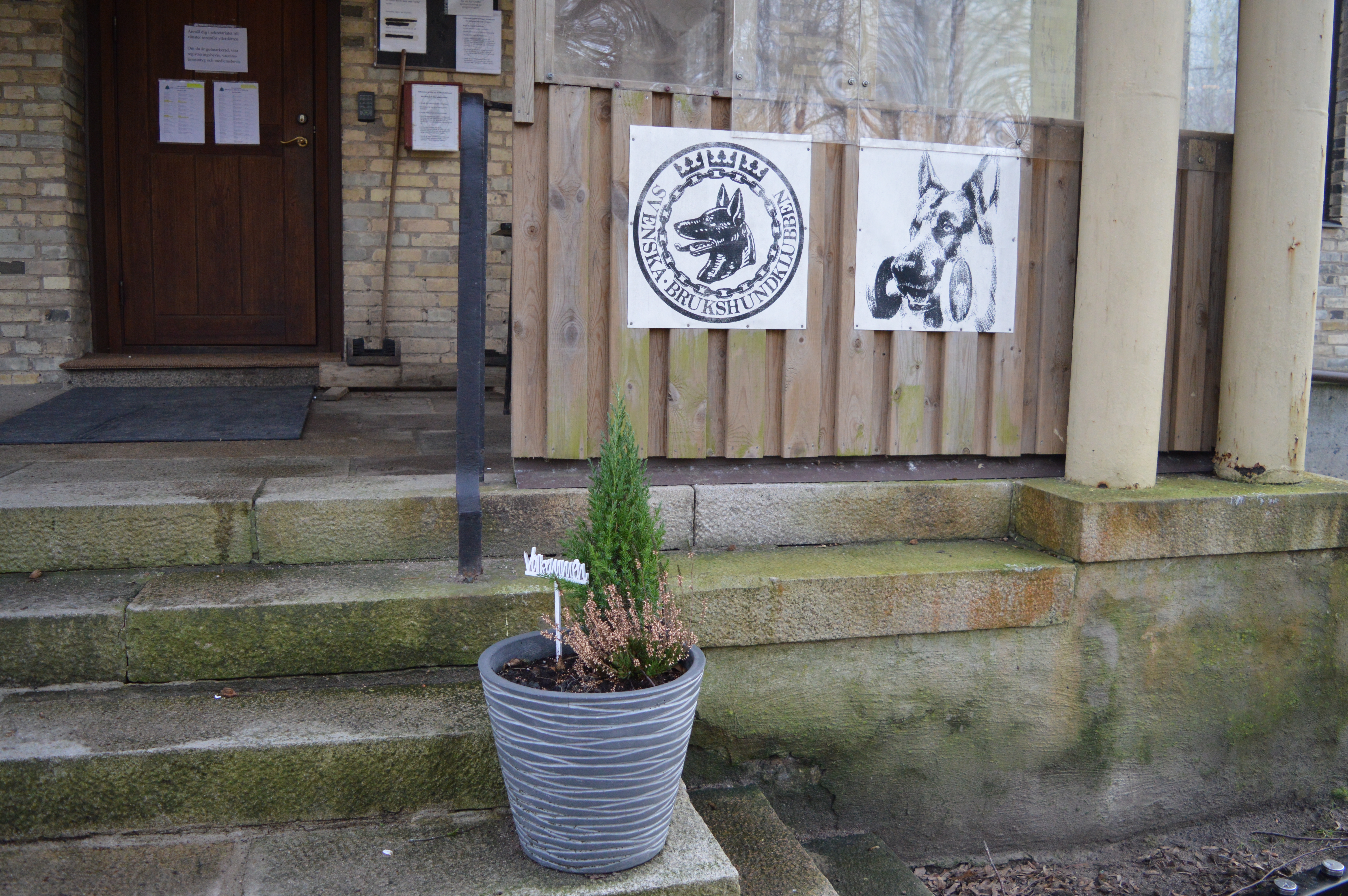 Bild på klubbstugans entre med en blomkruka med en välkommenskylt,