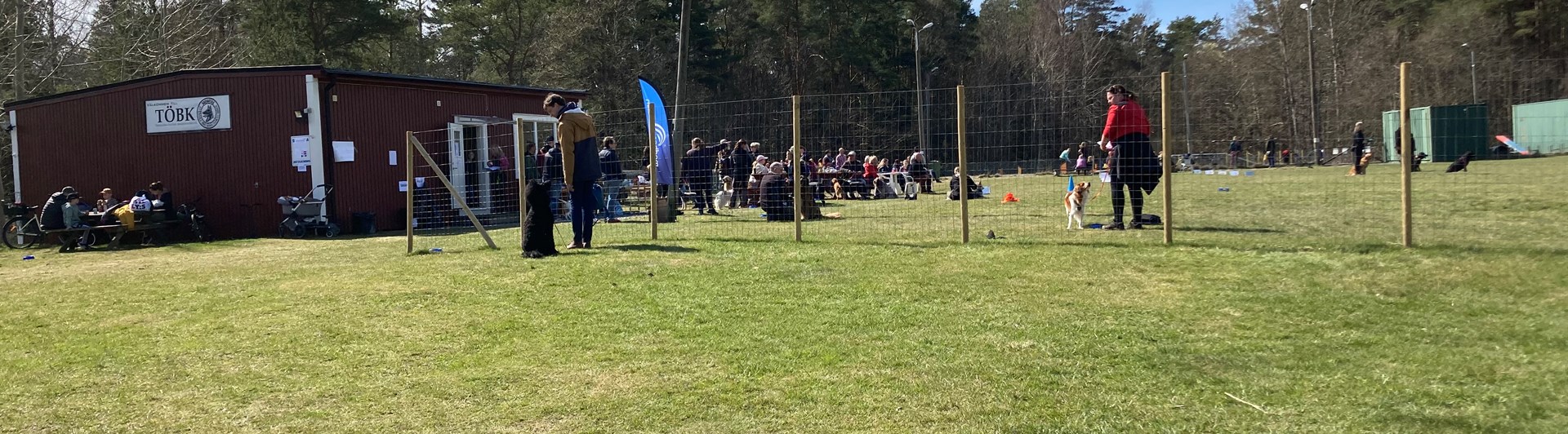 Röd klubbstuga med gräsplan framför. Många människor och hundar.