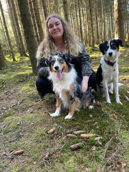 Tjej som sitter på huk bakom en hund som sitter framför henne. Bredvid henne sitter en svart vit hund