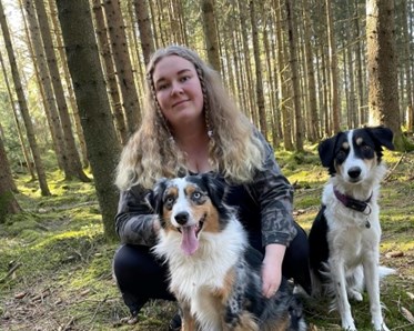 Tjej som sitter på huk bakom en hund som sitter framför henne. Bredvid henne sitter en svart vit hund
