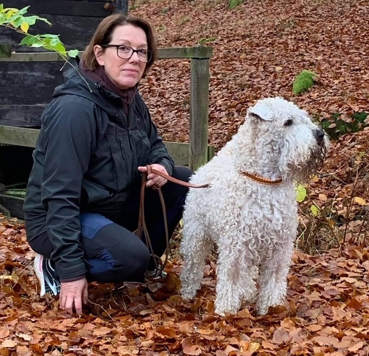 Vår instruktör Estelle Palmquist i svart jacka sitter på huk bland bruna höstlöv intill sin wheatenterrier.