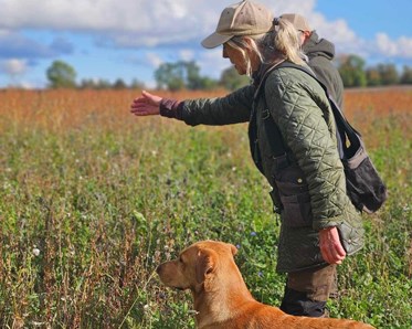 Else-Marie med blont hår, keps och grön jacka står i ett fält och anvisar riktning för sin gula labrador som står intill henne.