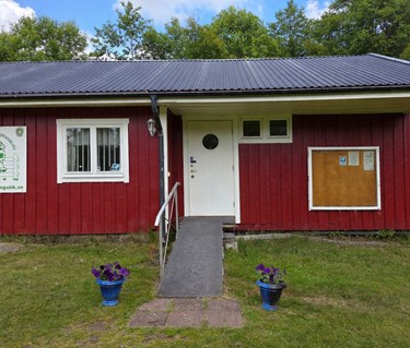 Röd klubbstuga med vita knutar och vit dörr. Blommor är placerade framför den handikappanpassade entrén.