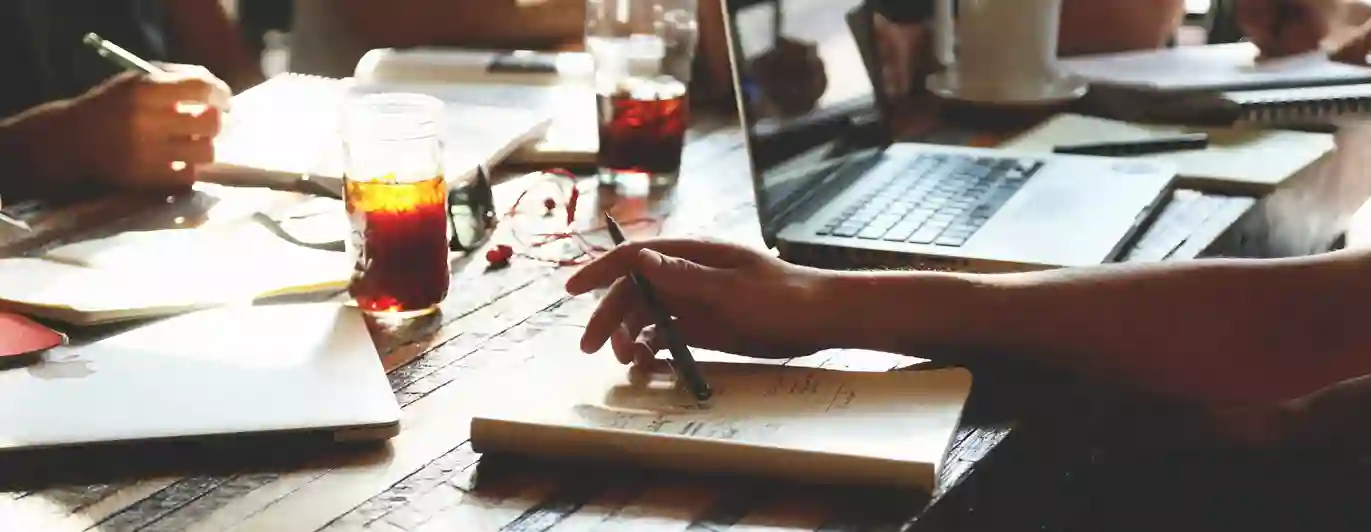 Bild på mötesbord med pennor, block, dator och skrivande händer.