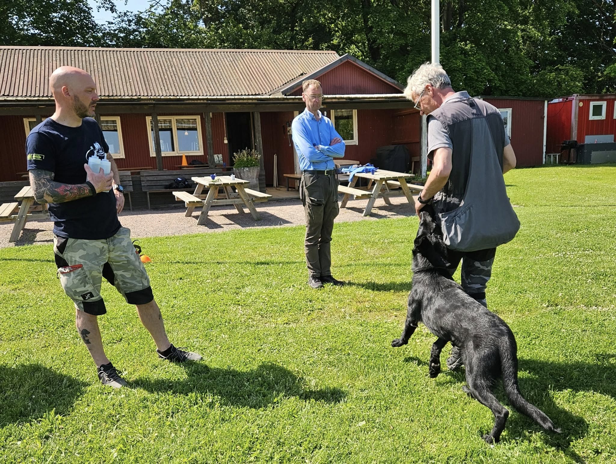 Svart schäfer med hundförare får instruktioner av instruktör som står bredvid. 