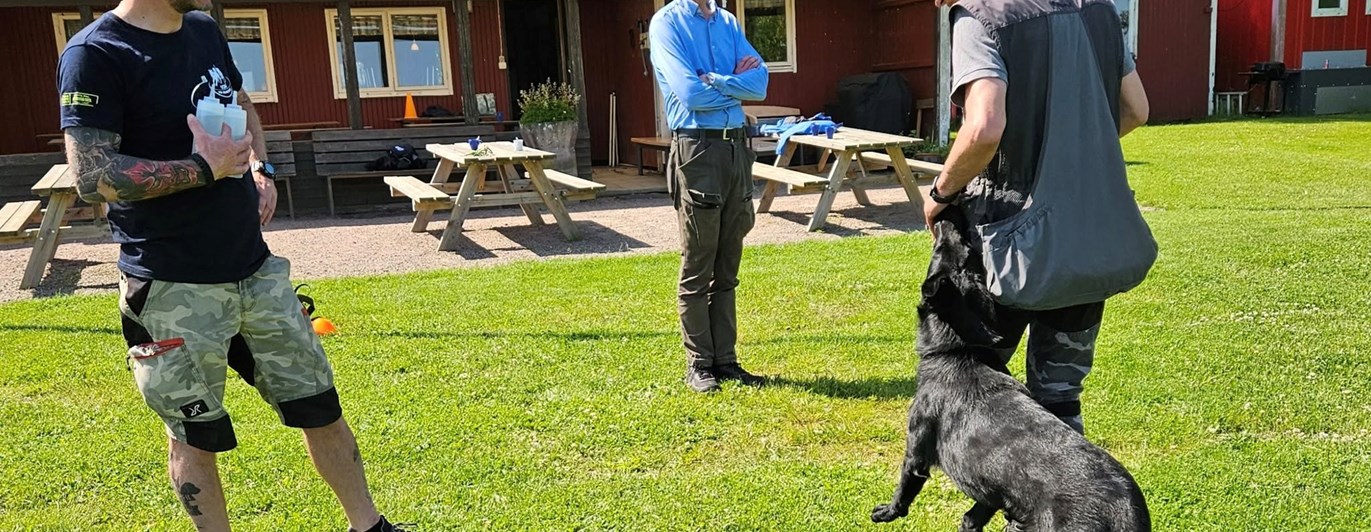 Svart schäfer med hundförare får instruktioner av instruktör som står bredvid.