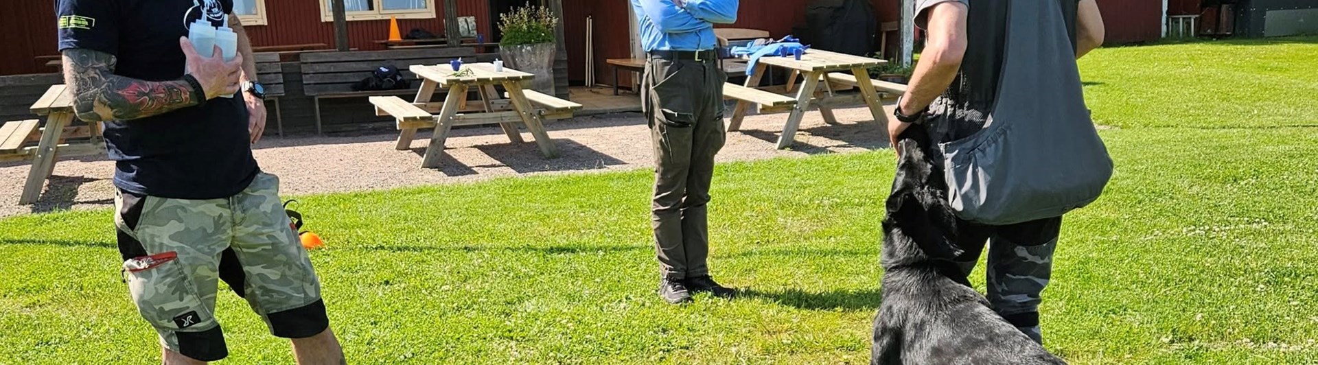 Svart schäfer med hundförare får instruktioner av instruktör som står bredvid.