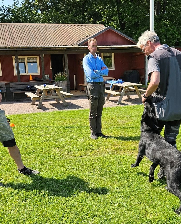 Svart schäfer med hundförare får instruktioner av instruktör som står bredvid.