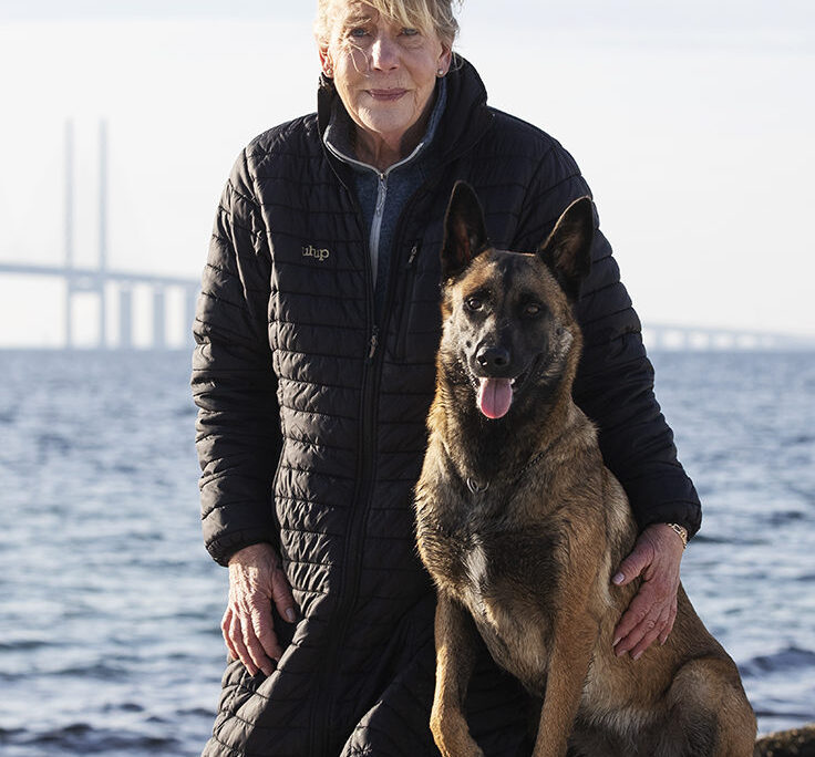Vår instruktör Lislotte Andersson i svart jacka står bredvid sin malinois med Öresundsbron i bakgrunden.