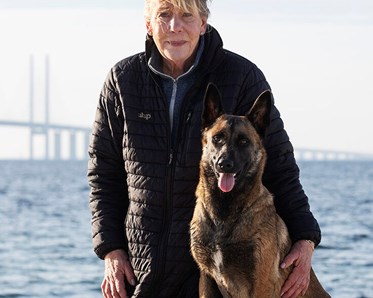 Vår instruktör Lislotte Andersson i svart jacka står bredvid sin malinois med Öresundsbron i bakgrunden.