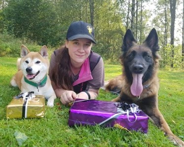 Kvinna som ligger på mage på gräset. Hon har keps på sig. bredvid henne på varsin sida ligger två hundar. Framför de ligger det ett lila paket