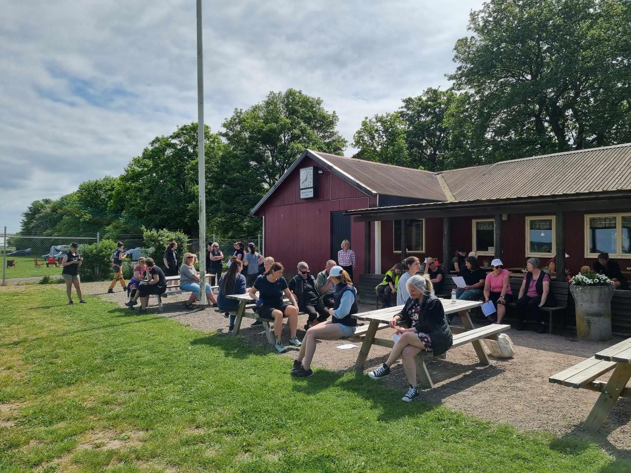 Grupp människor som fikar utomhus vid en röd stuga.