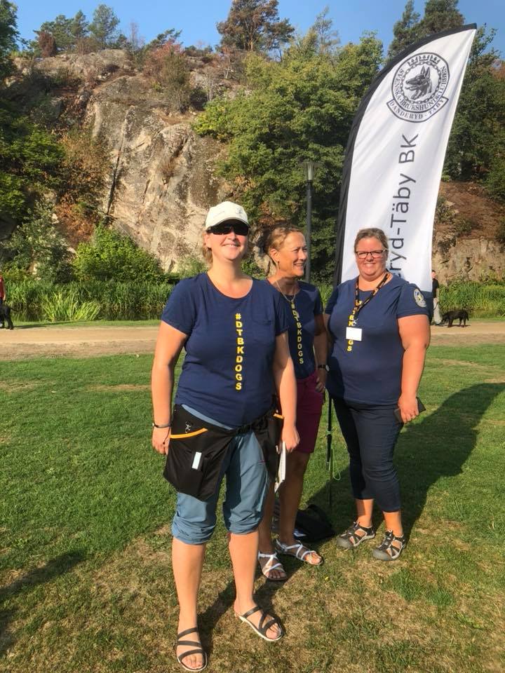 tre personer ler mot kameran med Danderyd-Täby brukshundklubbs flagga i bakgrunden