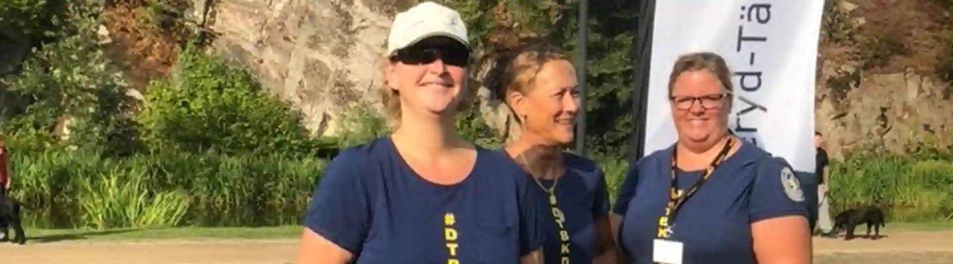 tre personer ler mot kameran med Danderyd-Täby brukshundklubbs flagga i bakgrunden