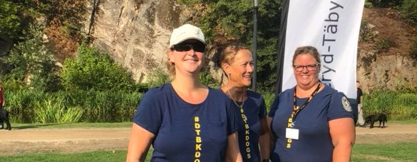 tre personer ler mot kameran med Danderyd-Täby brukshundklubbs flagga i bakgrunden