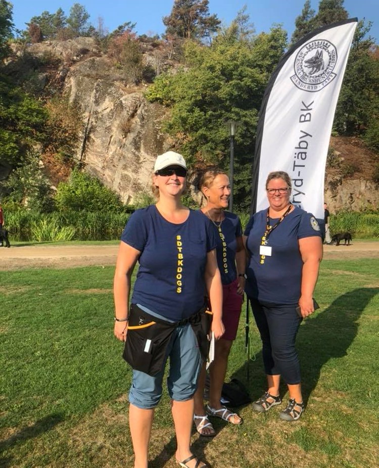 tre personer ler mot kameran med Danderyd-Täby brukshundklubbs flagga i bakgrunden