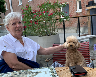 Yvonne i vit blus sitter utomhus vid ett träbord och klappar ljusbruna pudeln Alice som sitter på stolen intill.