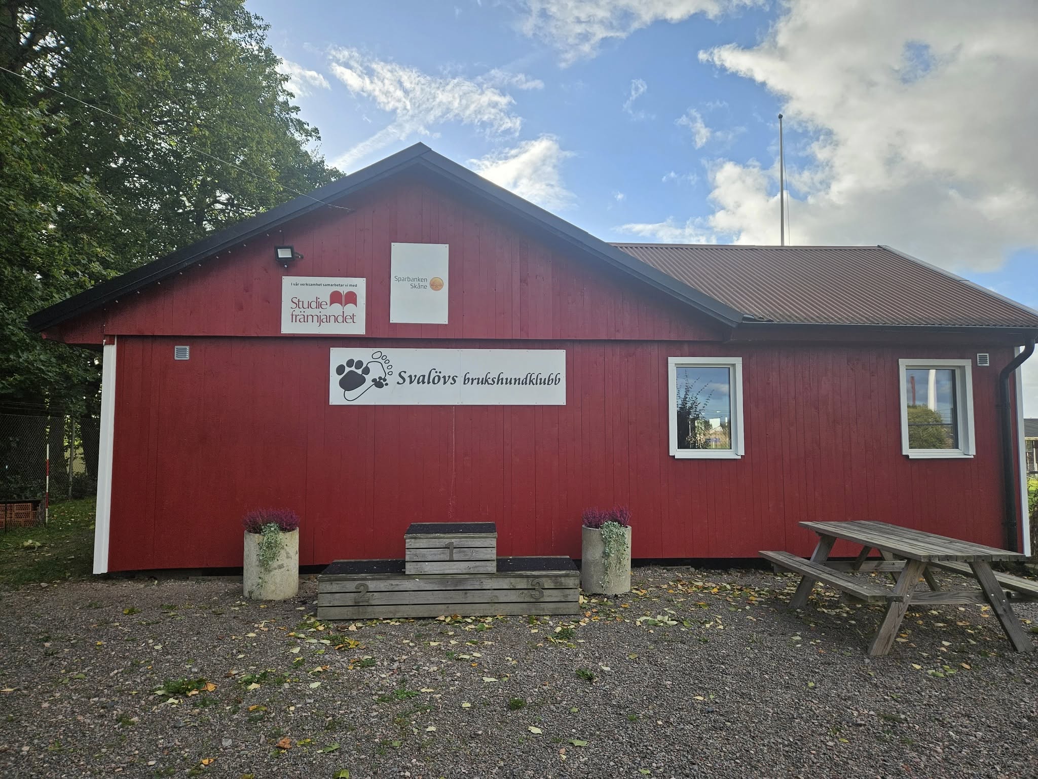 Röd klubbstuga i trä. På bilden syns också fina krukor med blommor. 