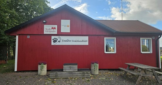 Röd klubbstuga i trä. På bilden syns också fina krukor med blommor.