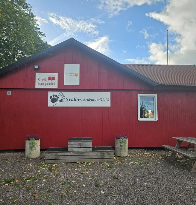Röd klubbstuga i trä. På bilden syns också fina krukor med blommor.