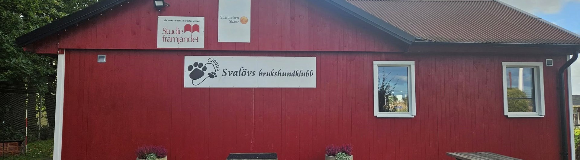 Röd klubbstuga i trä. På bilden syns också fina krukor med blommor.