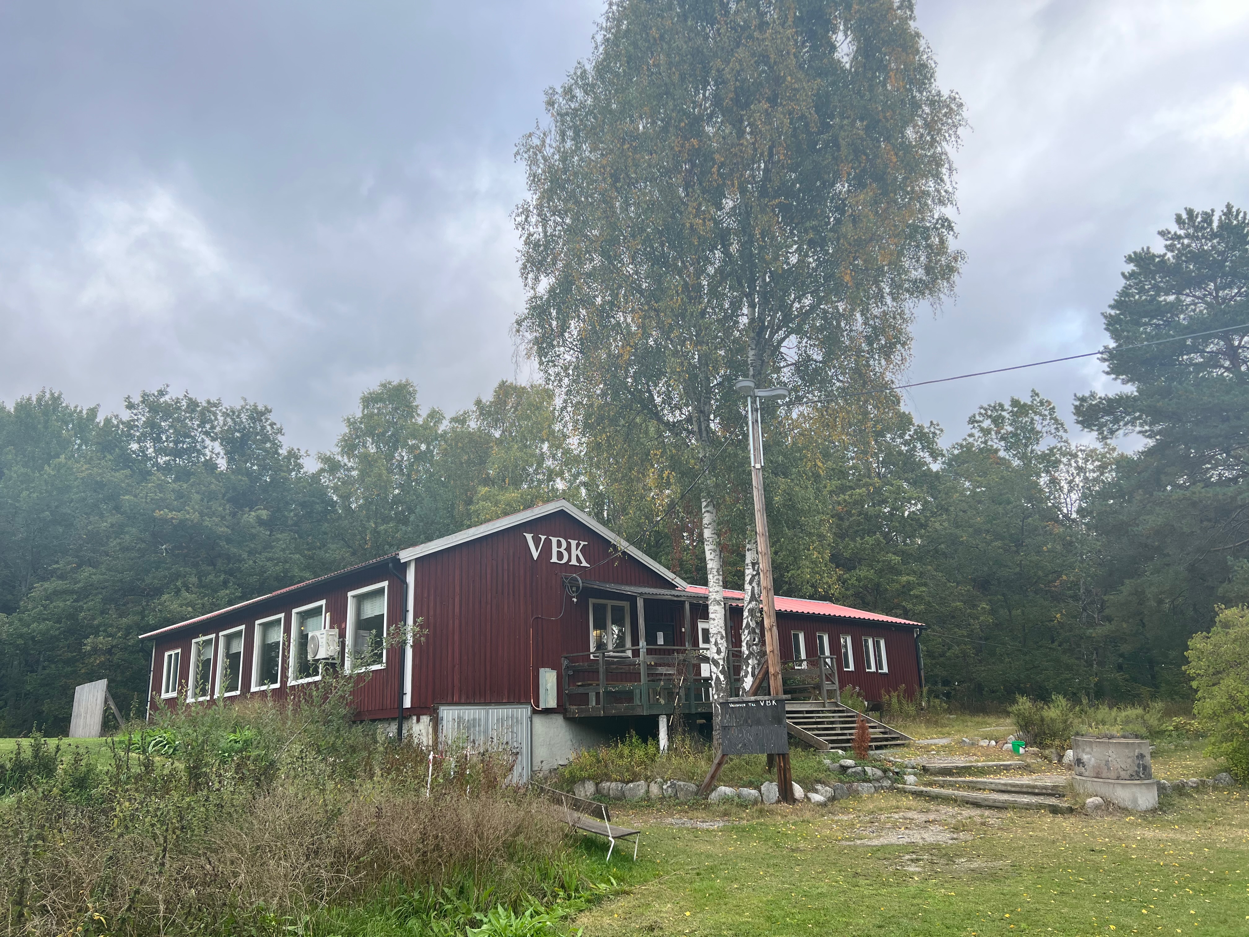 Rött hus med texten "VBK" i vitt. Framför huset finns en gräsyta och en stor björk.