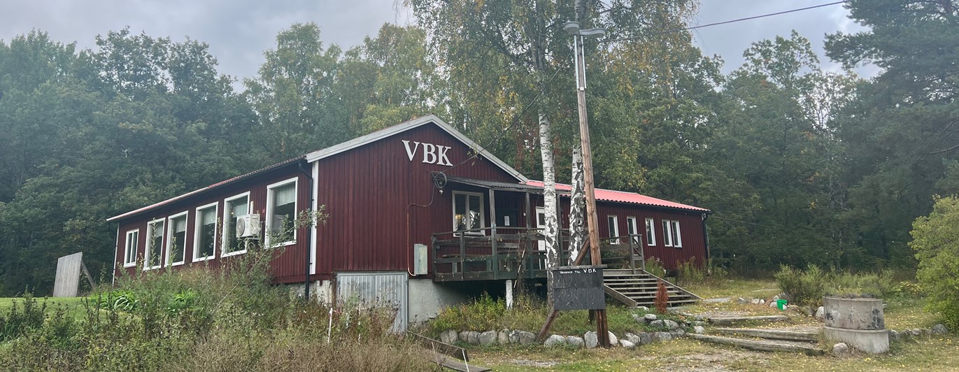Rött hus med texten "VBK" i vitt. Framför huset finns en gräsyta och en stor björk.