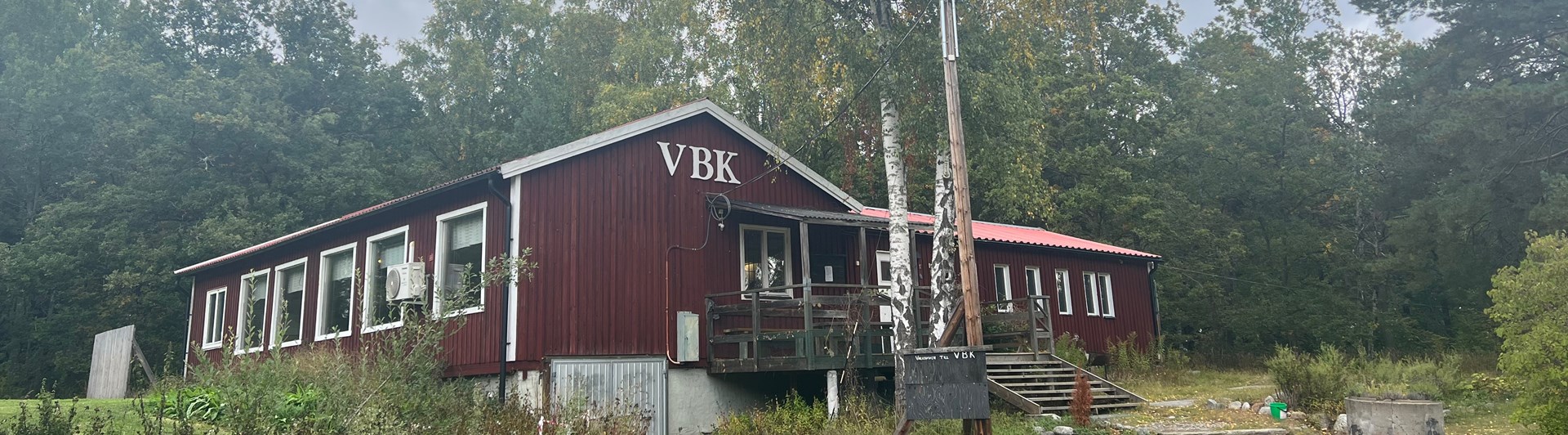Rött hus med texten "VBK" i vitt. Framför huset finns en gräsyta och en stor björk.
