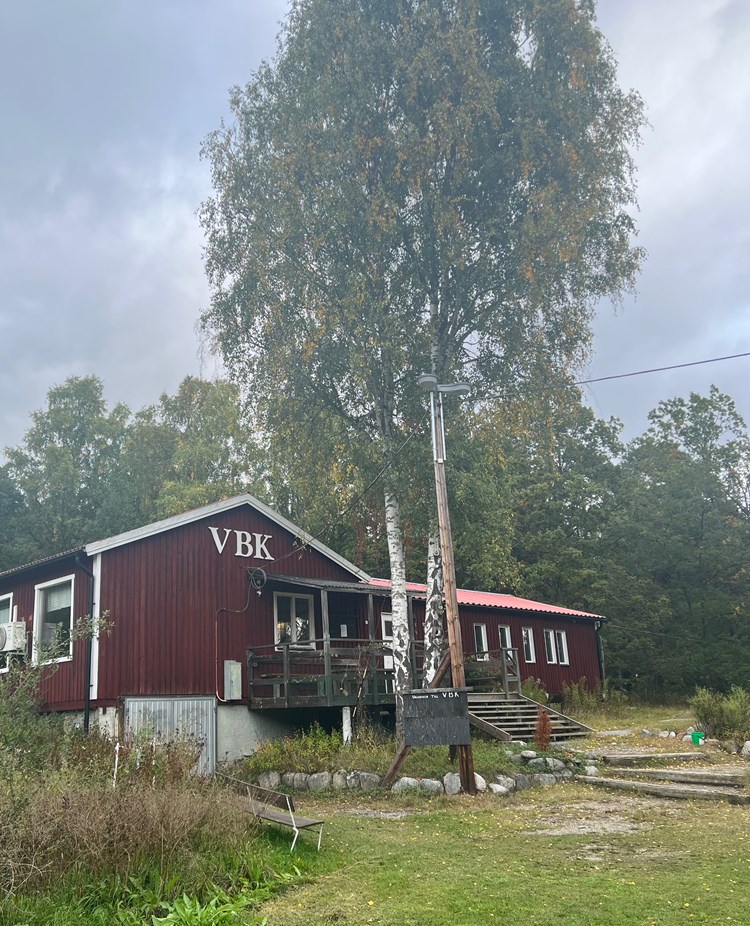 Rött hus med texten "VBK" i vitt. Framför huset finns en gräsyta och en stor björk.