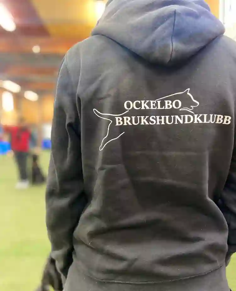 Ryggtavla på en person som bär en svart hoodie med Ockelbo brukshundklubbs logga. I bakgrunden syns ett ekipage som tränar kontakt med hunden sittandes vid sidan om föraren. 