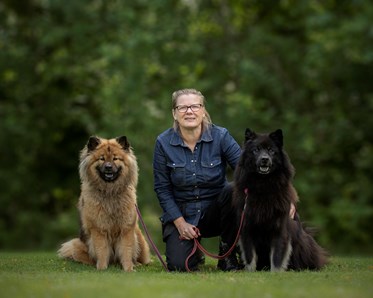 Förare som sitter på knä med varsin hund vid sin sida. En brun Eurasier på ena sidan och en svart Eurasier på andra sidan.