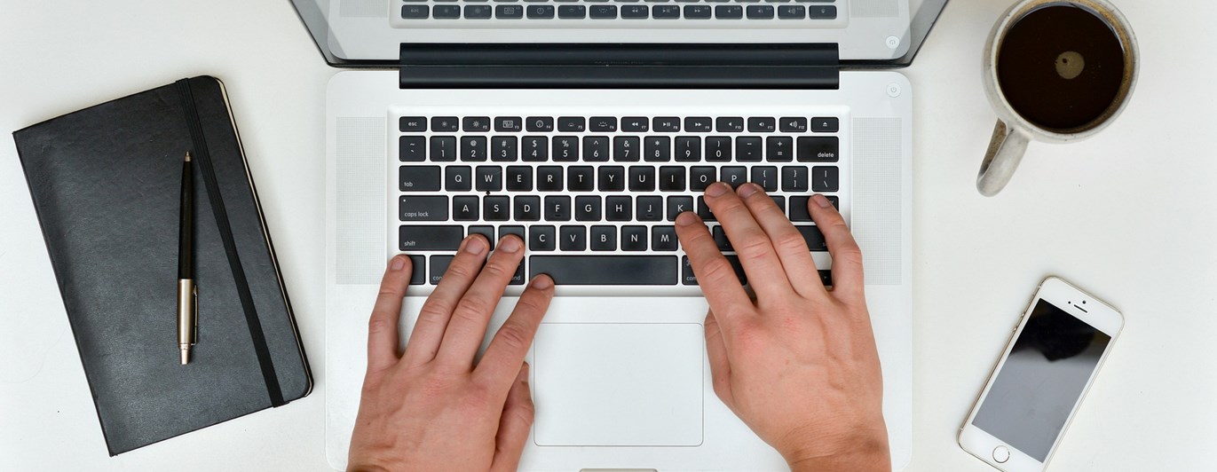 Bild som visar skrivande händer på en bärbar dator, anteckningsbok, mobil och kaffemugg.