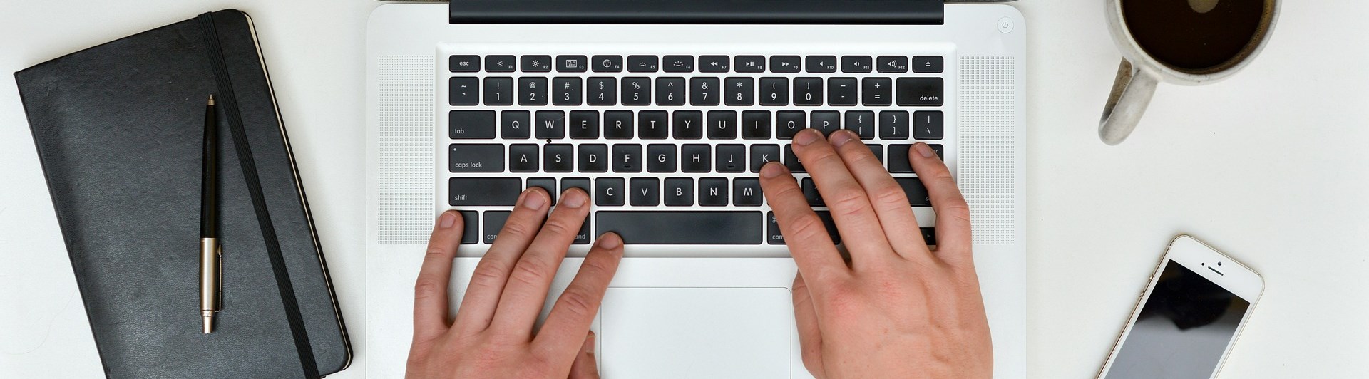 Bild som visar skrivande händer på en bärbar dator, anteckningsbok, mobil och kaffemugg.