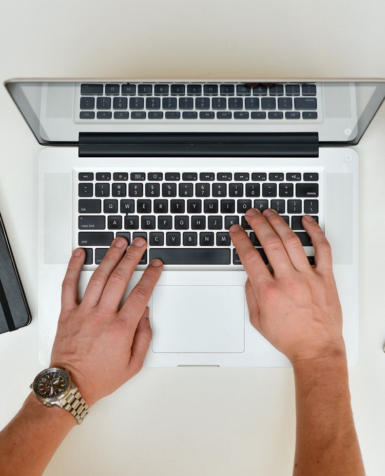 Bild som visar skrivande händer på en bärbar dator, anteckningsbok, mobil och kaffemugg.