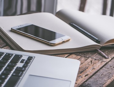 Bild som visar en bärbar dator, mobil och anteckningsblock på ett träbord.