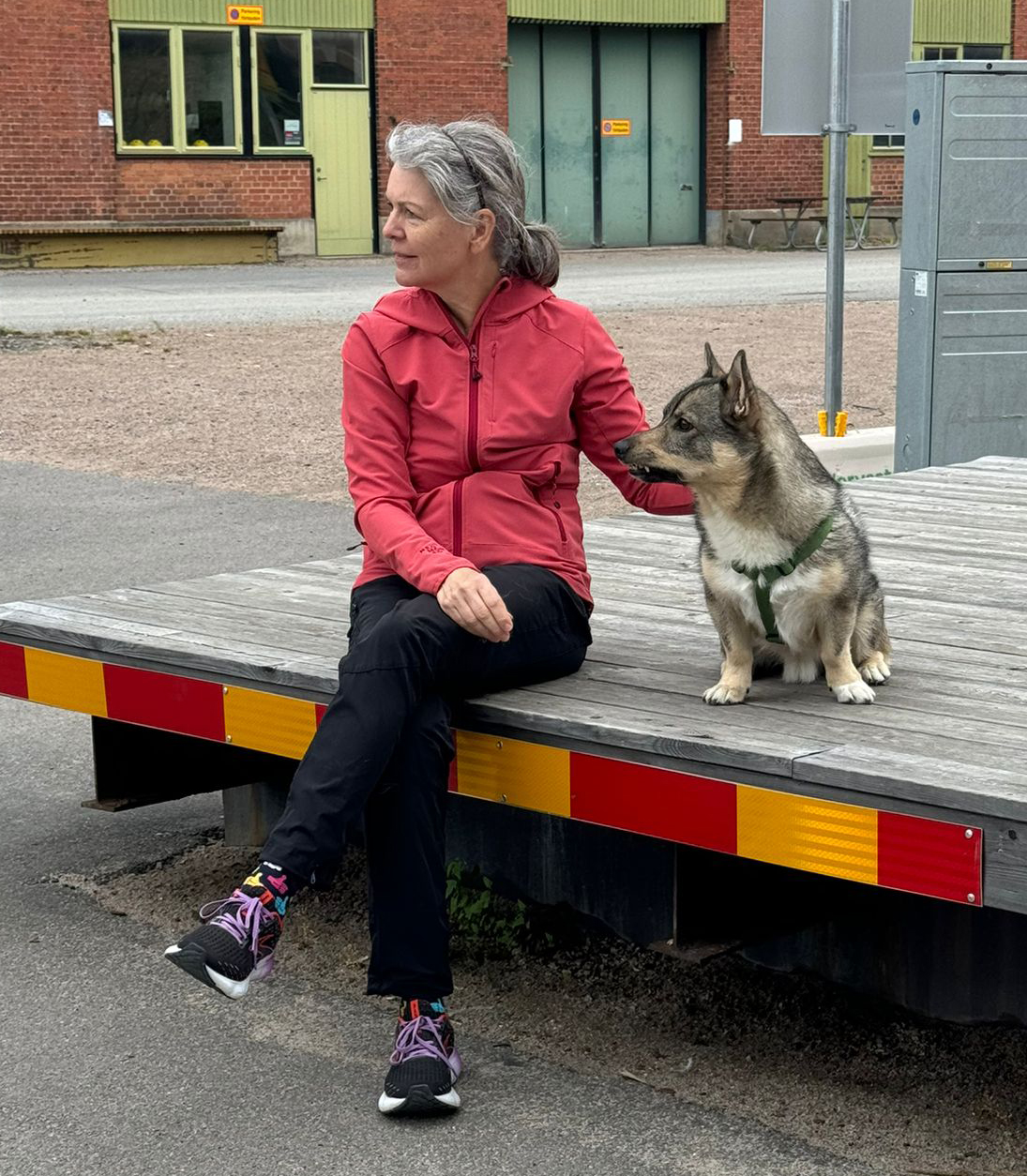 Raja Sundman i röd jacka sitter på en lastkaj med sin hund, en västgötaspets, bredvid sig.