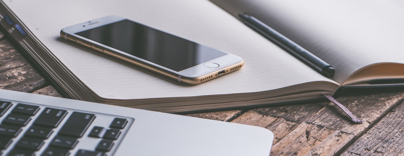 Bild på administrationsarbete med bärbardator, mobil och anteckningsblock.