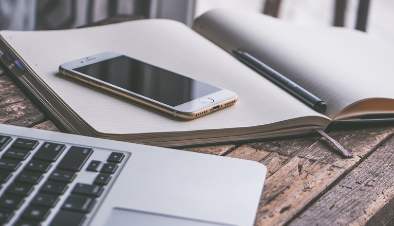 Bild på administrationsarbete med bärbardator, mobil och anteckningsblock.