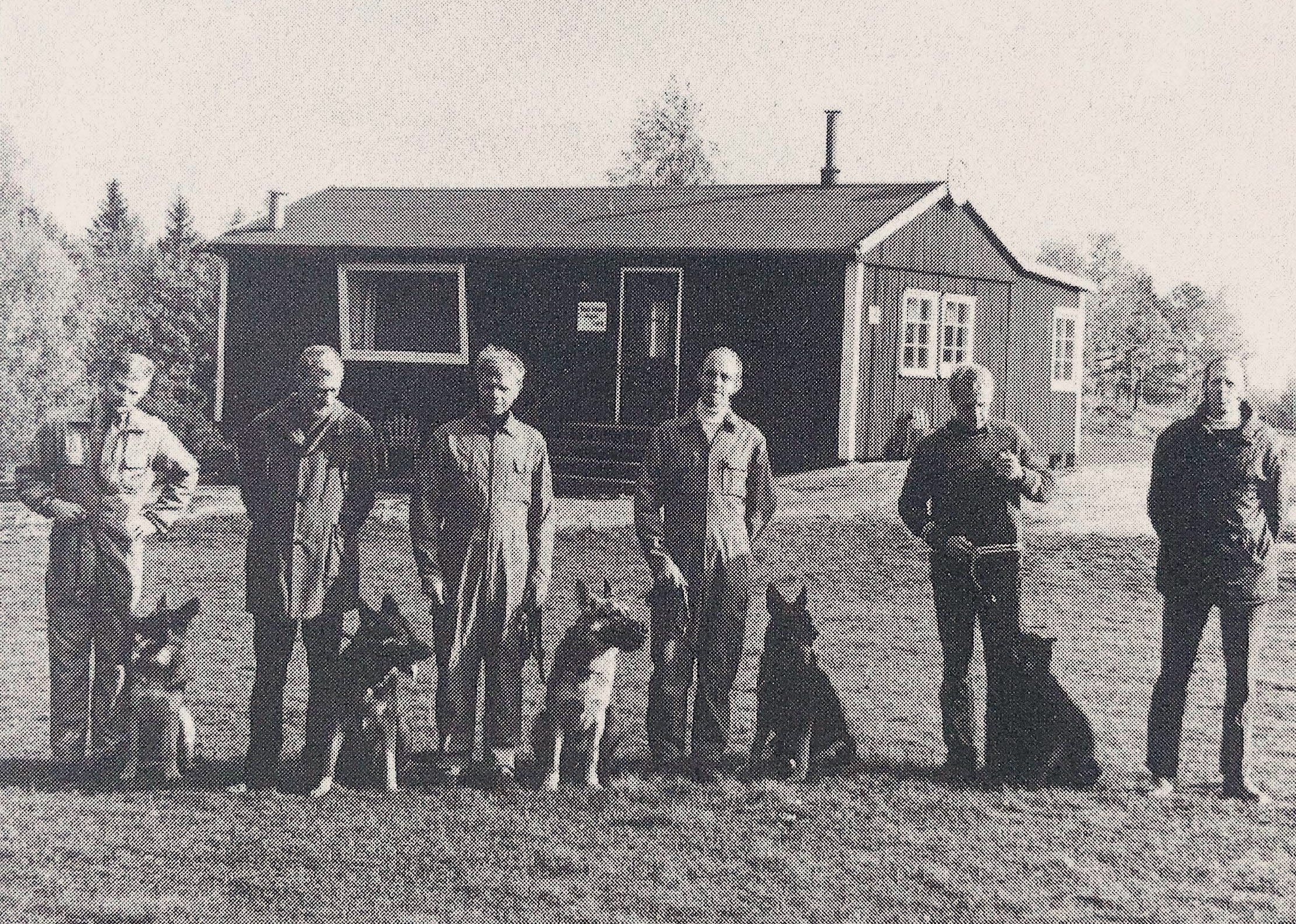 Äldre bild föreställande män med hundar framför klubbstuga